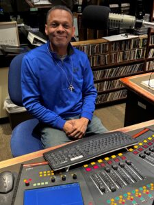 WSGE DJ Benjamil Guest - Wearing blue shirt and smiling at control panel.