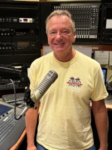 WSGE DJ Johnny B wearing yellow shirt, smiling, standing at microphone.