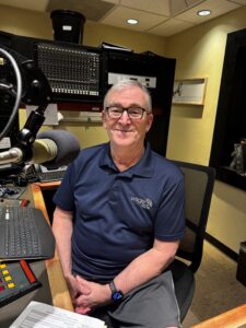 WSGE DJ Larry Strout wearing blue shirt and glasses, smiling in control room.