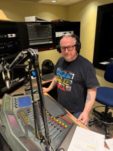 WSGE DJ Mark K - Wearing dark shirt and glasses, sitting at control panel.